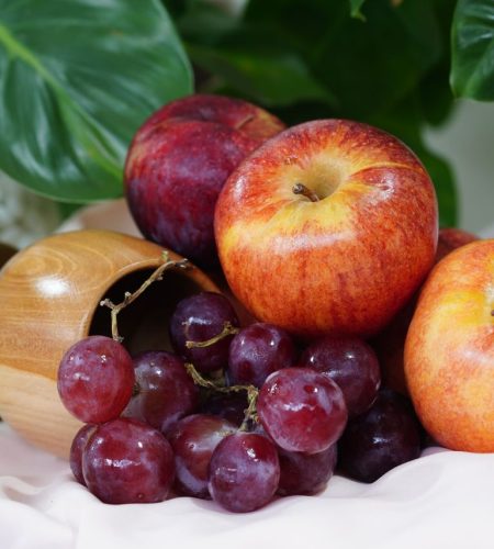 A still life of apples, grapes, and plums beside a wooden bowl and green leaves.
